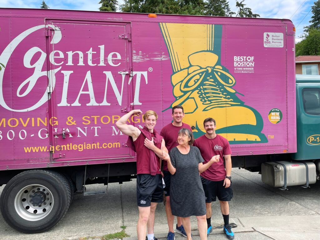 Three Gentle Giant movers dressed in maroon tshirts and black shorts are standing with a female customer in front of the purple Gentle Giant moving truck.