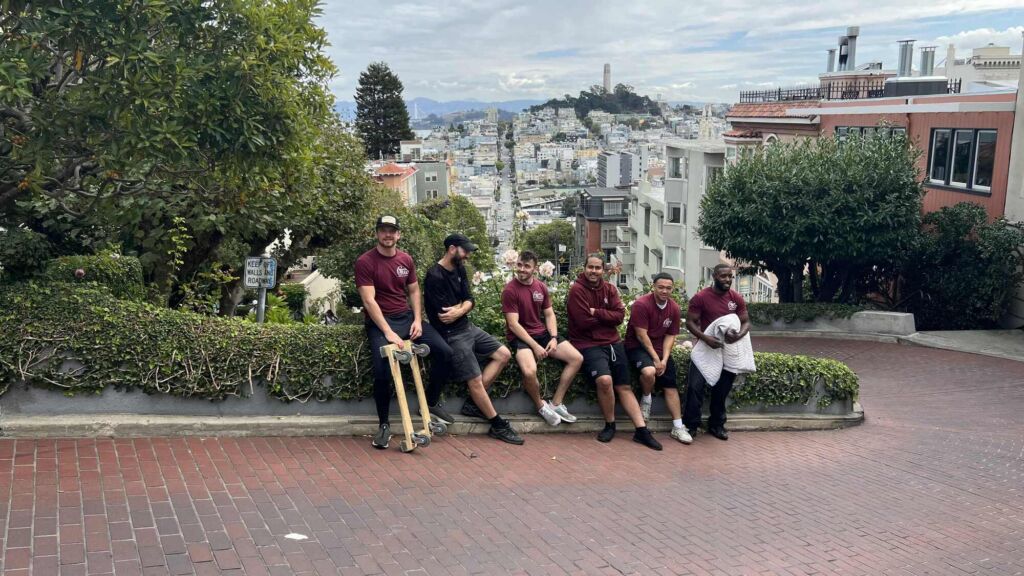 Six Gentle Giant movers are sitting on a wall on Lombard Street. The view of the city of San Francisco is behind them.