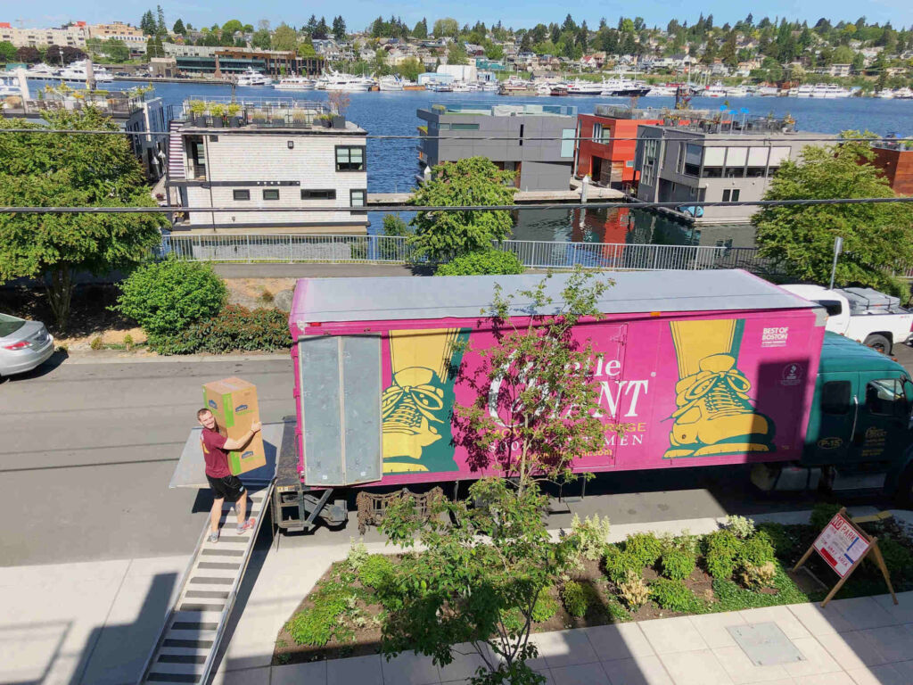 A Gentle Giant mover is walking up a ramp connected to a Gentle Giant moving truck. He his carrying two boxes. The purple moving truck is parked in front of a house. In the background, behind the moving truck, there are houses and a lake with boats on it.