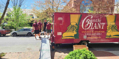 Four Gentle Giant movers are standing on the back of a purple and yellow moving truck. One mover is standing in front of them beside a metal ramp. The truck is parked in front of a brownstone church.