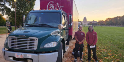 Two Gentle Giant Moving Company crew members stand smiling in front of a green Gentle Giant moving truck with the U.S. Capitol building visible in the background. The scene is set on a grassy area with fallen autumn leaves and a clear evening sky.