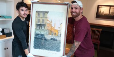 Two professional movers from Gentle Giant Moving Company carefully holding a framed artwork wrapped in protective material. Both movers are smiling and wearing gloves for safe handling in a well-lit, neatly arranged room.